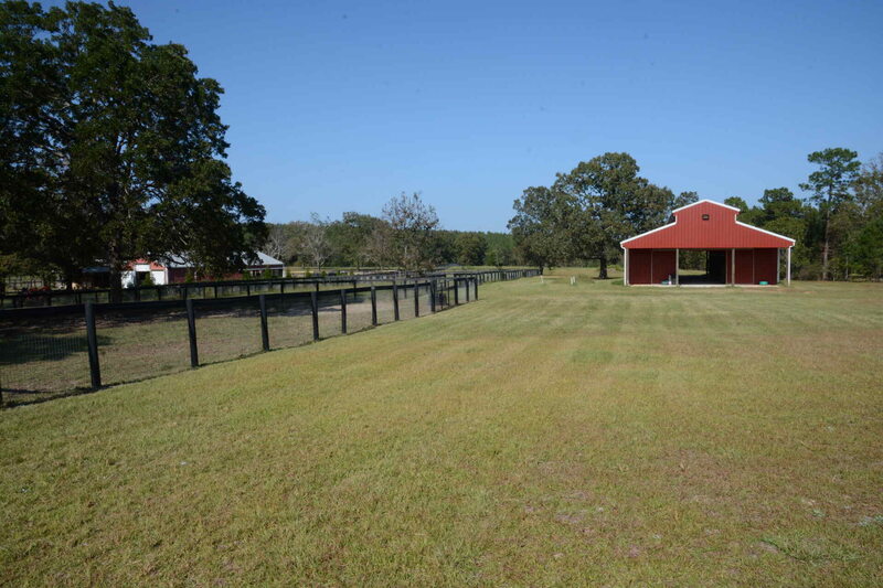 Red horse barn with open grassy area in front, black wooden fence to the left, and trees in the background.